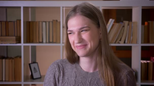 Крупный план портрета молодой привлекательной студентки, делающей смешное выражение лица и показывающей язык, смотрящей в камеру в библиотеке колледжа в помещении — стоковое видео
