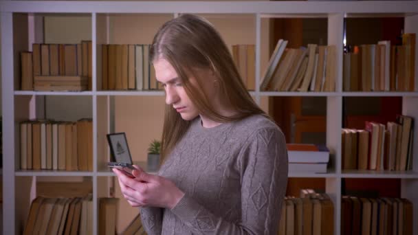 Крупный план портрета молодой привлекательной студентки, использующей телефон и смотрящей в камеру в библиотеке колледжа в помещении — стоковое видео