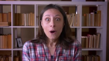 Genç çekici kız öğrencinin kapalı kütüphanede kameraya bakarak heyecanlı ve şaşırmış yakın çekim çekimi