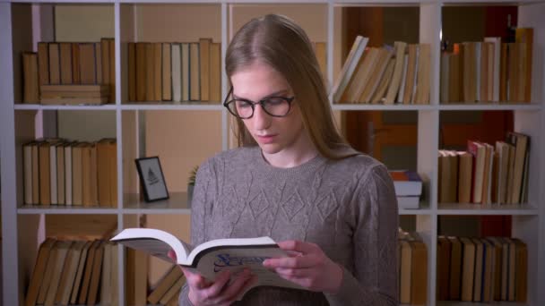 Retrato de cerca de una joven estudiante atractiva en gafas leyendo un libro y sonriendo mirando a la cámara en la biblioteca de la universidad en el interior — Vídeo de stock