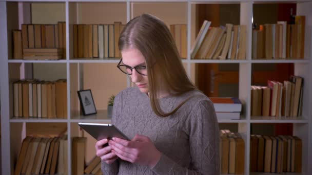 Gros plan portrait de jeune étudiante séduisante dans des lunettes à l'aide de la tablette et souriant en regardant la caméra dans la bibliothèque du collège à l'intérieur — Video