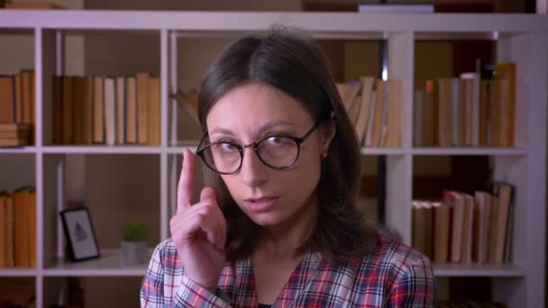 Close-up shoot van jonge aantrekkelijke vrouwelijke student die haar bril fixeer op camera met slimme uitdrukking in de bibliotheek binnenshuis — Stockvideo