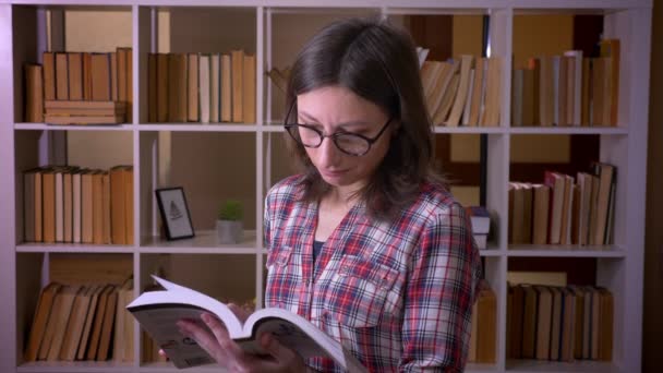 Närbild shoot av unga attraktiva kvinnliga student i glasögon läsa en bok leende tittar på kameran i biblioteket inomhus — Stockvideo