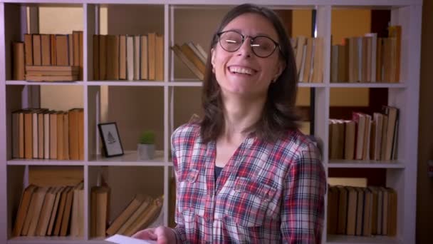 Gözlüklü genç çekici kız öğrencinin kapalı kütüphanede kameraya mutlu bir şekilde bakarak mutlu bir şekilde gülerek kitap okumasının yakın çekim çekimi — Stok video
