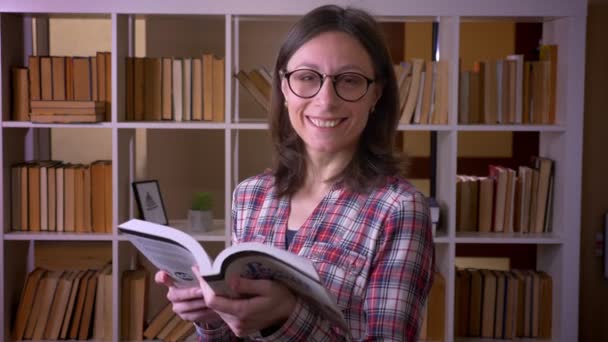 Zbliżenie strzelać z udany młody atrakcyjny żeński student w okulary czytanie a książka uśmiechnięty patrząc w kamera w The Biblioteka wewnątrz — Wideo stockowe