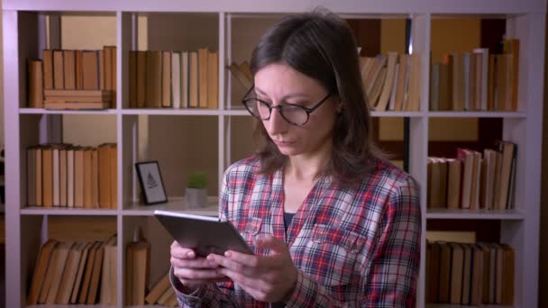 Tiro de close-up de jovem estudante atraente em óculos usando o tablet e olhando para a câmera na biblioteca dentro de casa — Vídeo de Stock
