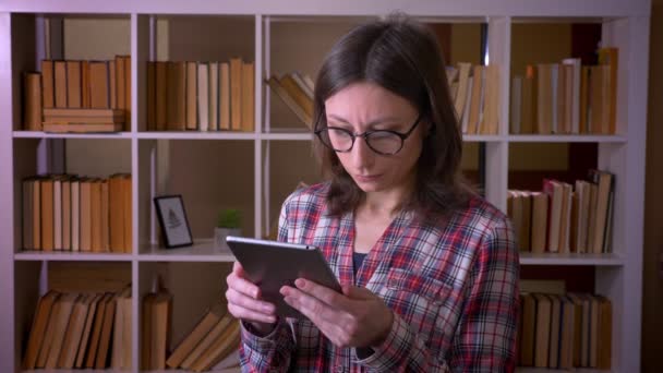 Střílejte mladé atraktivní studentky ve skleničkách pomocí tabletu a dívat se na kameru s úsměvem v univerzitní knihovně uvnitř — Stock video