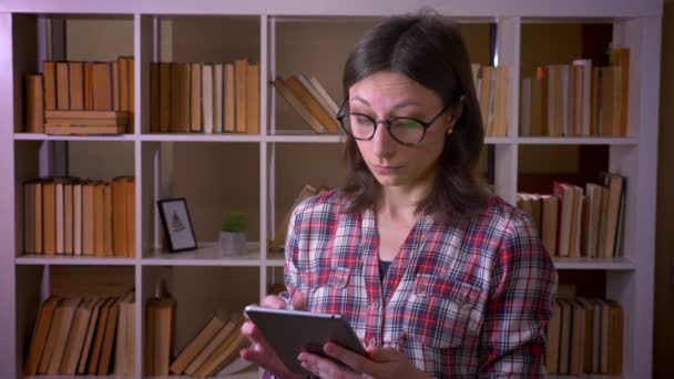 Tiro de close-up de jovem estudante atraente em óculos usando o tablet e mostrando tela verde para câmera na biblioteca dentro de casa — Vídeo de Stock