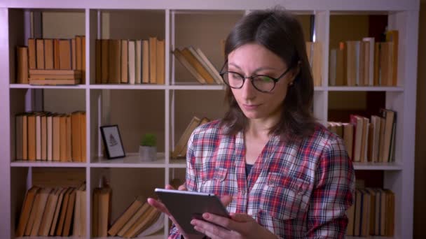 Zbliżenie strzelać młodej atrakcyjnej studentki w okularach za pomocą tabletu i pokazując zielony ekran chrominancji do kamery w bibliotece w pomieszczeniu — Wideo stockowe