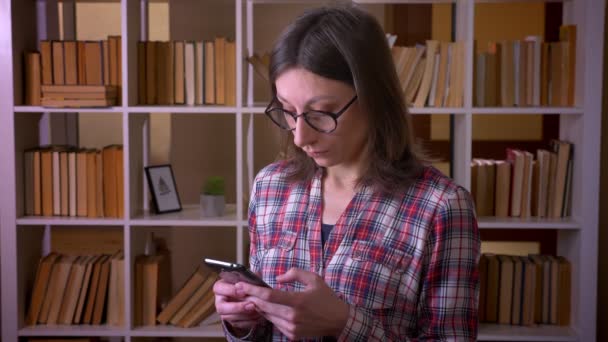 Gros plan de jeune étudiante séduisante dans des lunettes en utilisant le téléphone et en regardant la caméra dans la bibliothèque à l'intérieur — Video