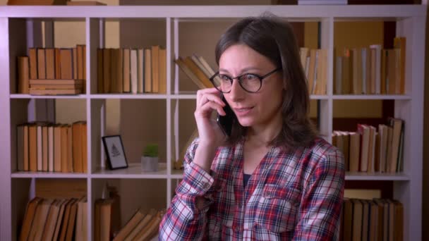 Gros plan de jeune étudiante séduisante dans des lunettes ayant un appel téléphonique dans la bibliothèque à l'intérieur — Video