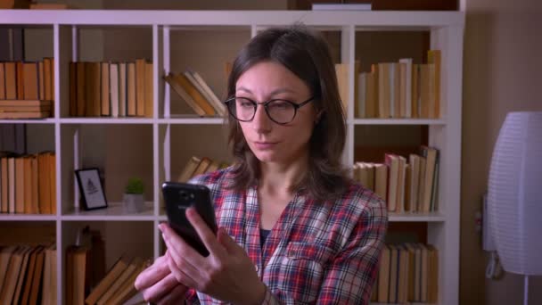 Primo piano ripresa di giovane studentessa attraente in occhiali avendo una videochiamata al telefono in biblioteca al chiuso — Video Stock