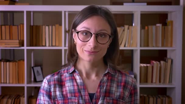 Close-up schieten van jonge aantrekkelijke vrouwelijke student in glazen met een duim omhoog kijken naar camera in de bibliotheek binnenshuis — Stockvideo