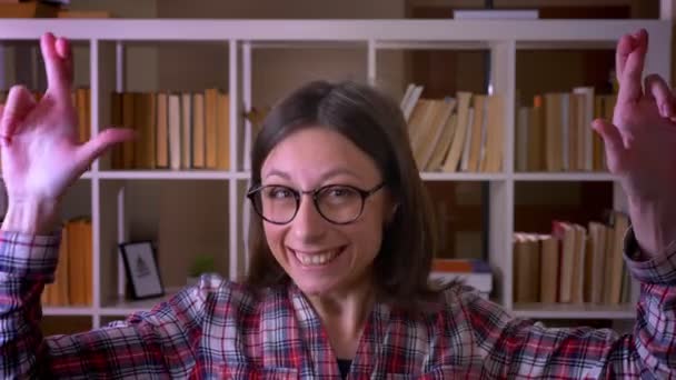 Close-up shoot van jonge aantrekkelijke vrouwelijke student met haar vingers gekruide glimlachend met hoop naar camera in de bibliotheek binnenshuis — Stockvideo
