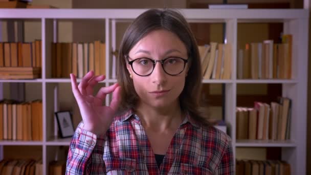 Gros plan de jeune étudiante séduisante dans des lunettes montrant signe correct regardant la caméra dans la bibliothèque à l'intérieur — Video