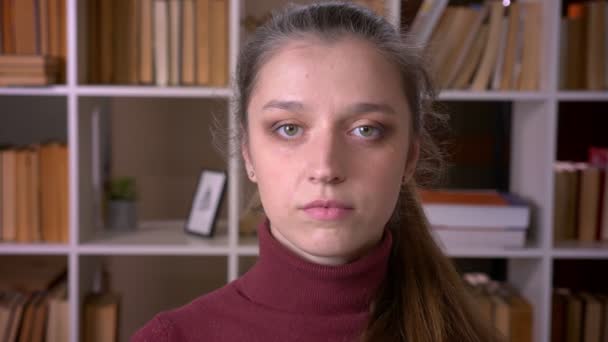 Retrato de cerca de una joven estudiante caucásica mirando a la cámara en la biblioteca de la unversidad en el interior — Vídeos de Stock