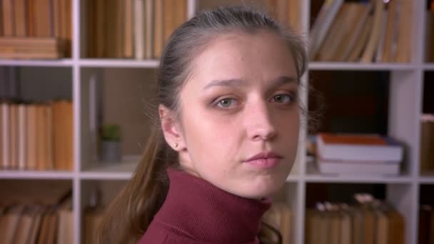 Retrato de primer plano de una joven estudiante caucásica girando y mirando la cámara en la biblioteca universitaria en el interior — Vídeos de Stock