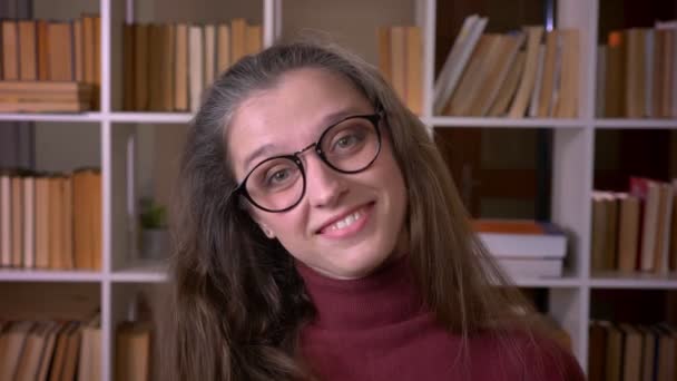 Närbild porträtt av unga kaukasiska kvinnlig student i glasögon leende med spänning tittar på kameran i kollegiet biblioteket inomhus — Stockvideo