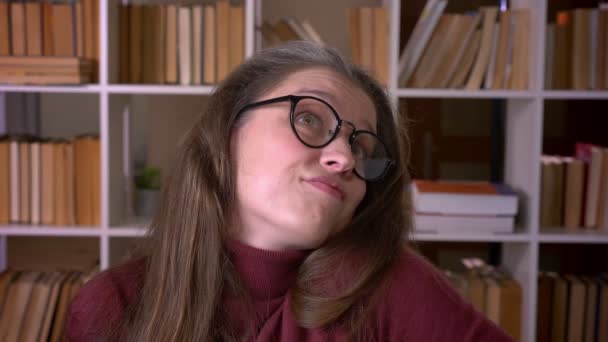 Closeup portrait of young caucasian female student in glasses smiling with confusion looking at camera in the college library indoors — Stock Video