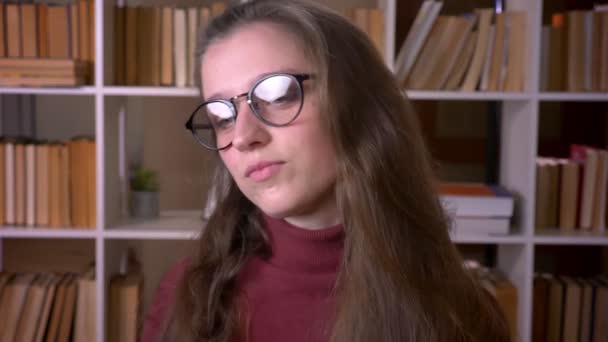 Retrato de cerca de una joven estudiante caucásica en gafas bailando felizmente delante de la cámara en la biblioteca universitaria en el interior — Vídeo de stock