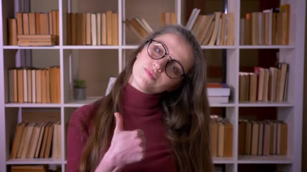 Крупный план портрета юной кавказской студентки в очках, показывающий большой палец, улыбающийся, счастливо смотрящий в камеру в библиотеке колледжа в помещении — стоковое видео