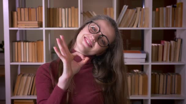 Retrato de primer plano de una joven estudiante caucásica con gafas que muestran un signo de aprobación sonriendo felizmente mirando a la cámara en la biblioteca universitaria en el interior — Vídeo de stock