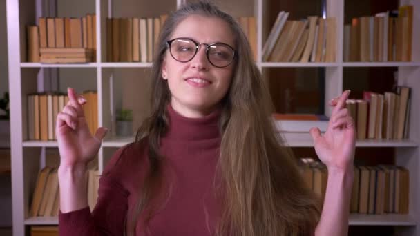 Nahaufnahme Porträt einer jungen kaukasischen Studentin mit Brille, die voller Hoffnung die Daumen drückt und in die Kamera in der Universitätsbibliothek blickt — Stockvideo