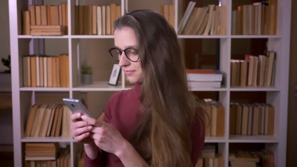 Gros plan portrait de jeune étudiante caucasienne dans des lunettes en utilisant le téléphone souriant regardant joyeusement la caméra dans la bibliothèque du collège à l'intérieur — Video