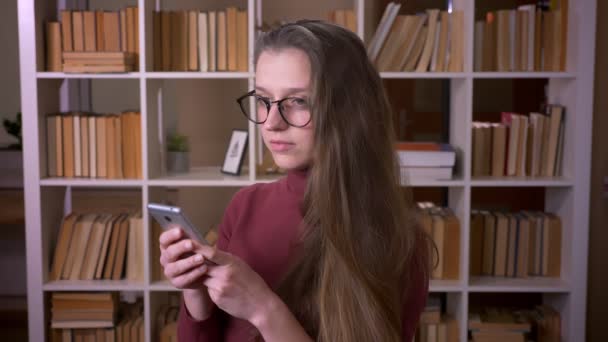 Closeup portrait of young caucasian female student in glasses browsing on the phone smiling happily looking at camera in the college library indoors — Stock Video