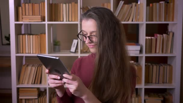 Zbliżenie portret młodego kaukaski studentka w okularach przy użyciu tabletu uśmiechając się i patrząc na kamerę w pomieszczeniu w bibliotece kolegium w pomieszczeniu — Wideo stockowe