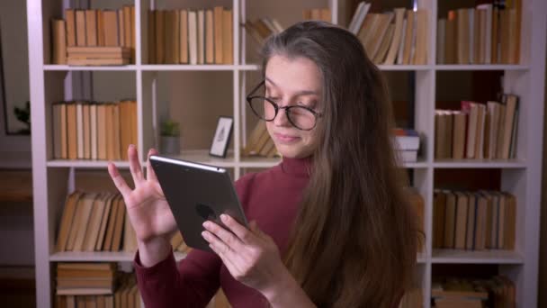 Κοντινό πλάνο της νεαρής καυκάσιας γυναίκας φοιτητής σε γυαλιά δακτυλογράφησης στο tablet σε εσωτερικούς χώρους στη βιβλιοθήκη κολλέγιο σε εσωτερικούς χώρους — Αρχείο Βίντεο