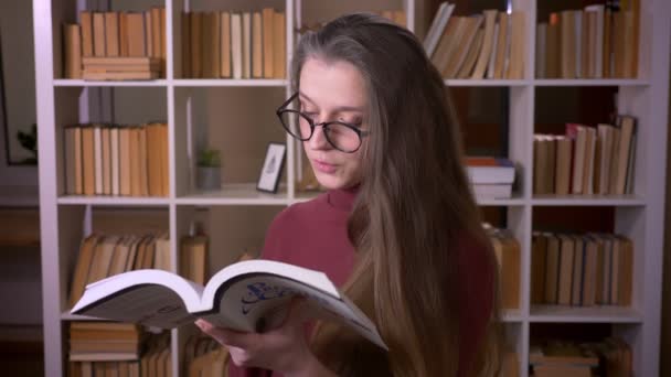 Retrato de close-up de jovem estudante branca em óculos lendo um livro e sorrindo alegremente olhando para a câmera na biblioteca da faculdade dentro de casa — Vídeo de Stock