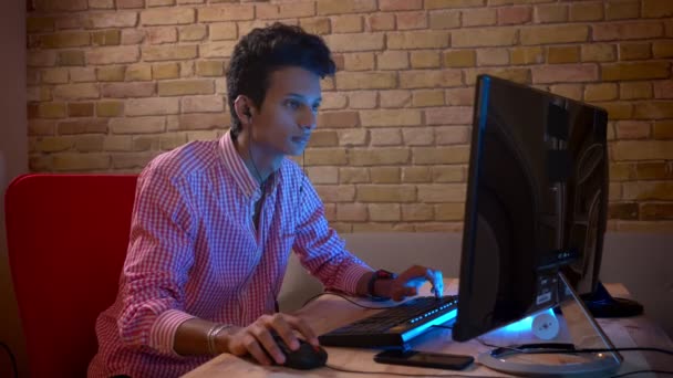 Primer plano de brote de joven indio atractivo macho en vibraciones jugando videojuegos en el ordenador interior en el acogedor apartamento con luz de neón — Vídeo de stock