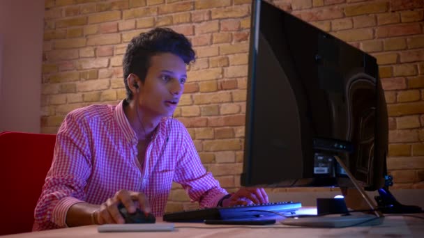 Zbliżenie strzelać młodego indyjskiego atrakcyjny mężczyzna w wibracje grając w gry wideo na mocy obliczeniowej i obchodzi szczęśliwie wewnątrz w przytulnym mieszkaniu z neonowym światłem — Wideo stockowe