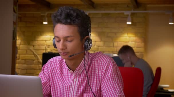 Primer plano de rodaje de indio joven hombre de negocios en los auriculares que tienen una llamada utilizando el ordenador portátil en la oficina en el interior — Vídeos de Stock