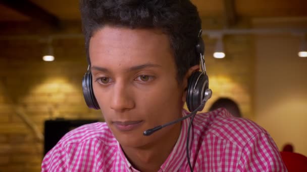Primer plano de la sesión de indio joven empleado en los auriculares que tienen una llamada utilizando el ordenador portátil en la oficina en el interior — Vídeos de Stock