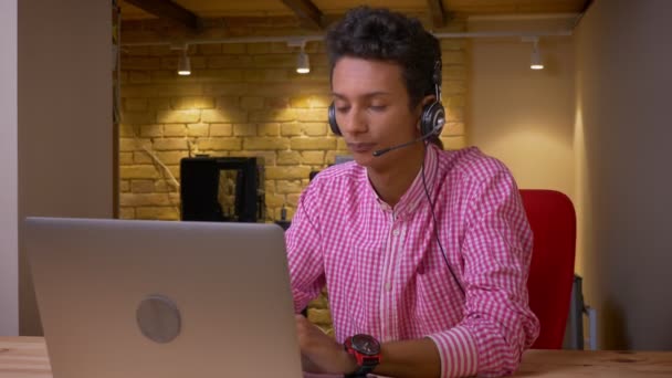 Primo piano ripresa di indiano giovane dipendente in cuffie avere una chiamata utilizzando il computer portatile sul posto di lavoro in ufficio al chiuso — Video Stock