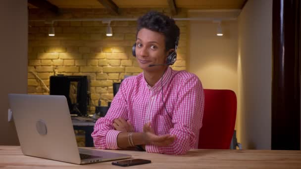 印度年轻商人在耳机的特写镜头看着相机微笑，在办公室的室内笔记本电脑 — 图库视频影像