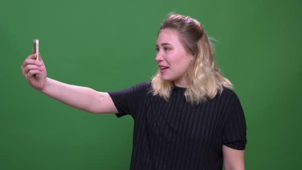 Closeup portrait of young pretty blonde caucasian female having a video call on the phone with background isolated on green — Stock Video