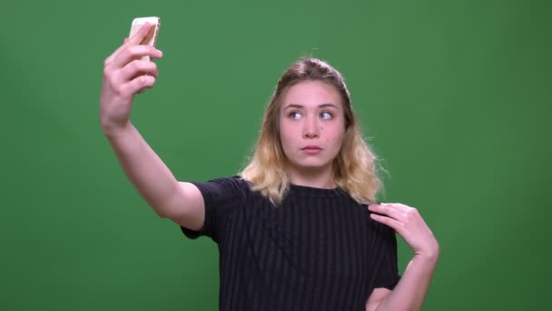 緑の上に隔離された背景を持つ携帯電話で自分撮りを取る若いかわいいブロンドの白人女性のクローズアップ肖像画 — ストック動画