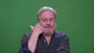 Gözlüklü kıdemli gri saçlı işadamı, yeşil renk arka planında düşünceli bir şekilde izole edilmiş sakalını kaşıyarak.