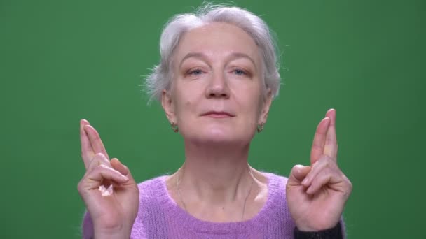 Grand-mère aux cheveux gris en pull violet prie avec les doigts croisés isolés sur fond vert chromatique . — Video