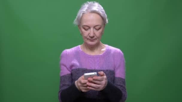 보라색 스웨터에 수석 회색 머리 여자 녹색 크로마키 배경에 고립 된 스마트 폰과 함께 작동. — 비디오