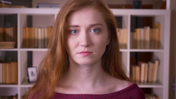 Крупный план портрета молодой рыжеволосой привлекательной кавказской студентки, смотрящей на камеру в библиотеке колледжа в помещении — стоковое видео