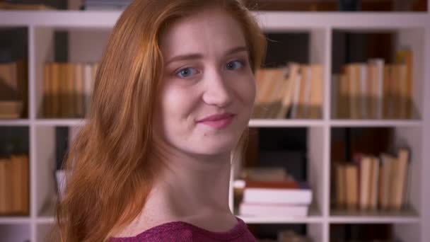 Closeup portrait of young redhead attractive caucasian female student turning and looking at camera smiling happily in the college library — Stock Video