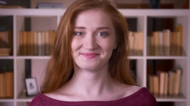 Close-up retrato de jovem ruiva atraente caucasiana estudante olhando para a câmera sorrindo alegremente na biblioteca da faculdade — Vídeo de Stock