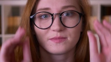 Üniversite kütüphanesinde utangaçlıkla gülümseyen kameraya bakan gözlüklü genç kızıl saçlı çekici beyaz kız öğrencinin yakın çekim portresi
