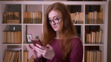 Üniversite kütüphanesinde telefonla mesaj atan gözlüklü genç kızıl saçlı çekici beyaz kız öğrencinin yakın çekim portresi