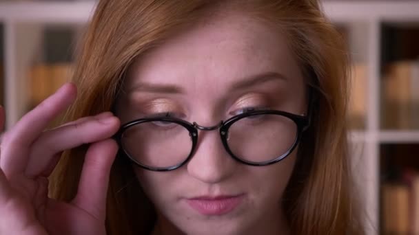 Closeup retrato de jovem ruiva atraente caucasiana estudante em óculos olhando para a câmera sorrindo alegremente na biblioteca da faculdade — Vídeo de Stock