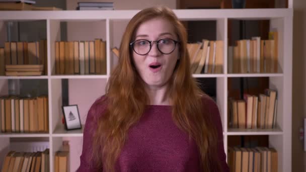 Крупним планом портрет молодої рудої привабливої білоруської студентки в окулярах збуджується і дивується, дивлячись на камеру в бібліотеці коледжу — стокове відео